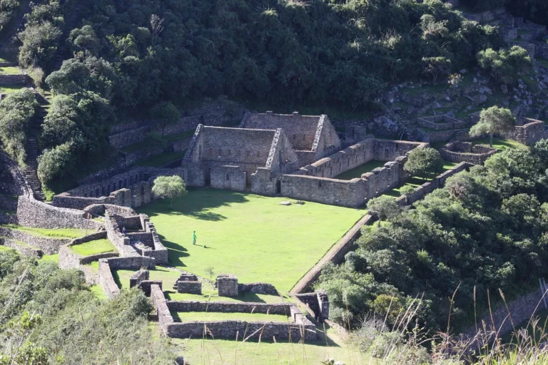 Praça principal de Choquequirao com gramado verde cercado por muros incas de pedra e mata fechada na encosta