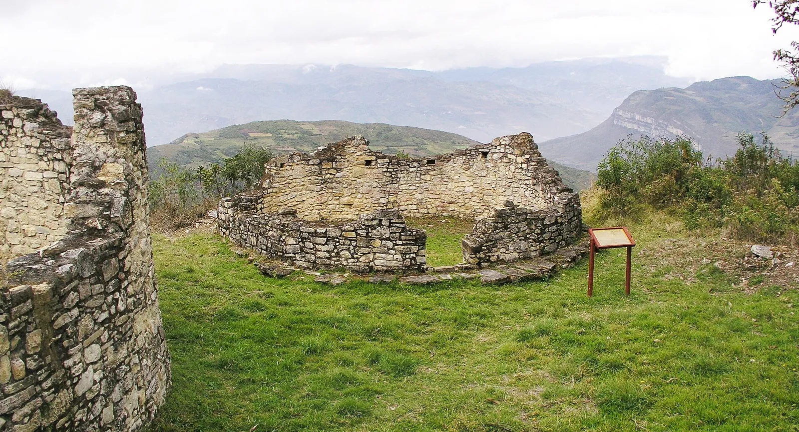 Ruínas de casas circulares da cultura Chachapoya dentro da llaqta de Kuélap, com placa informativa e montanhas ao fundo