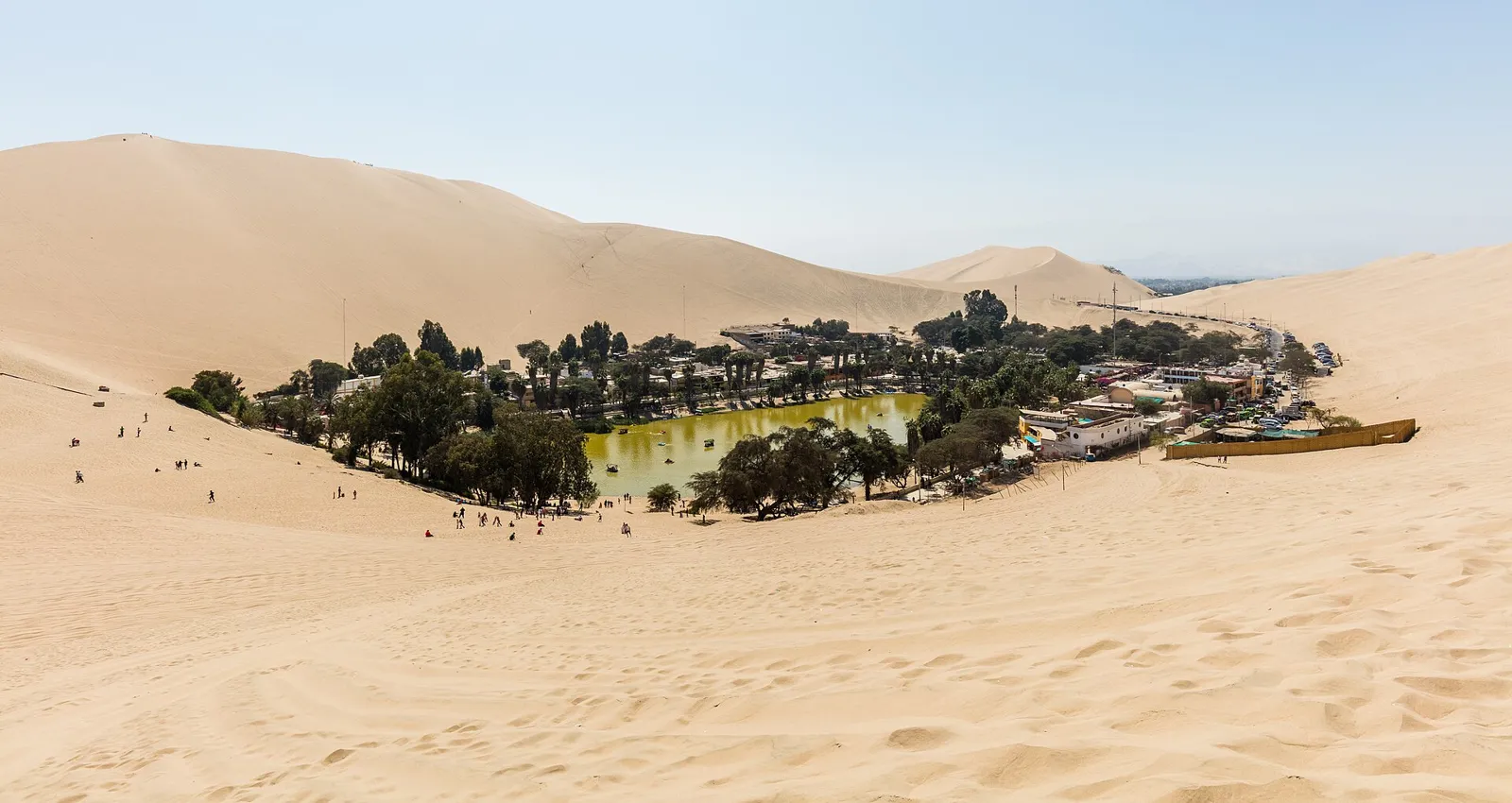 Lagoa do oásis de Huacachina cercada de palmeiras e construções, com grandes dunas de areia ao fundo, perto de Ica