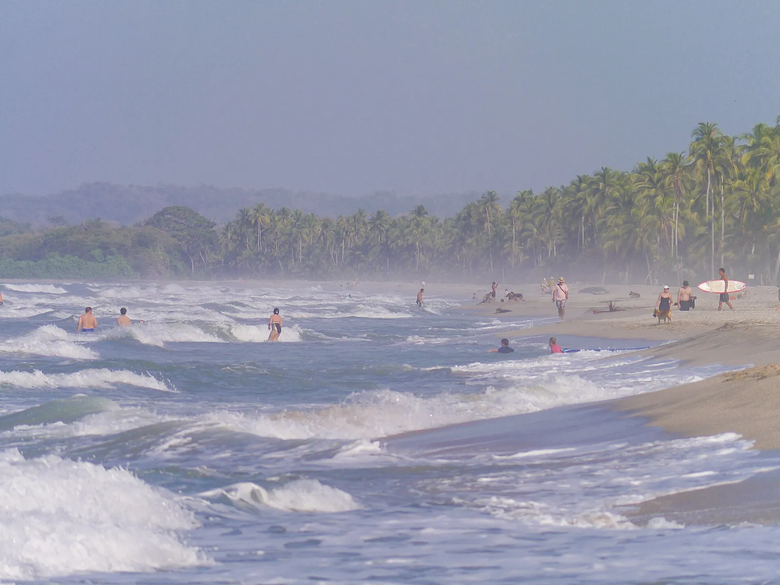 Praia de Palomino com ondas fortes, banhistas e surfista na areia, com coqueiros e montanhas da Sierra Nevada ao fundo